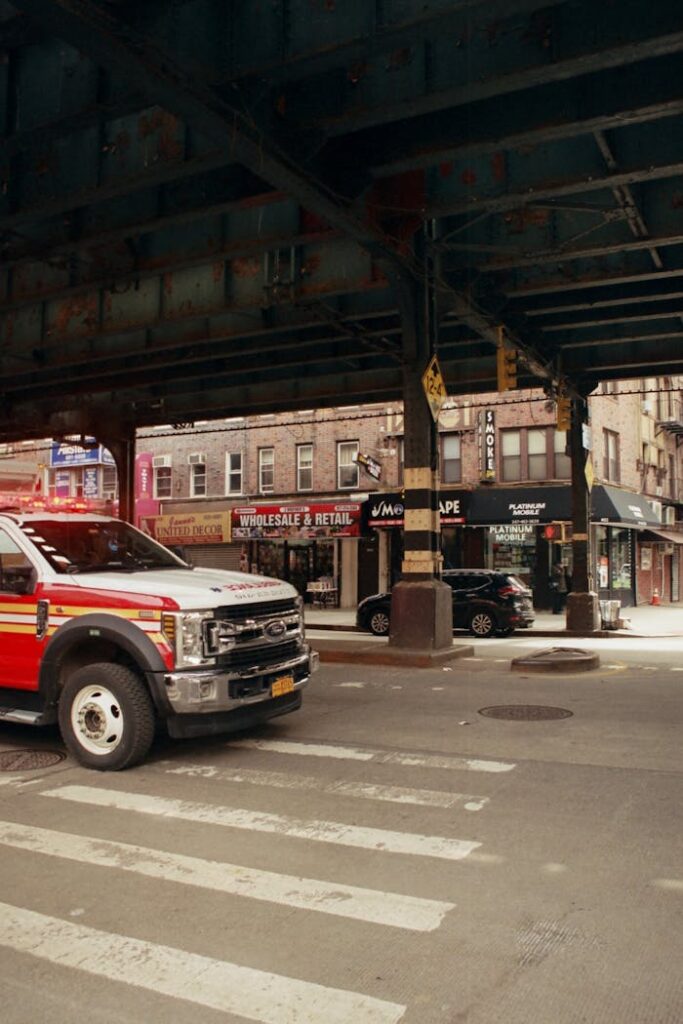 Red truck at city intersection with elevated road and shops in backdrop on a sunny day.
